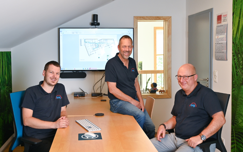 Drei Mitarbeiter der ibag Seedorf sitzen in einem Besprechungsraum an einem Tisch; im Hintergrund ist ein Monitor mit einem Gebäudeplan zu sehen.